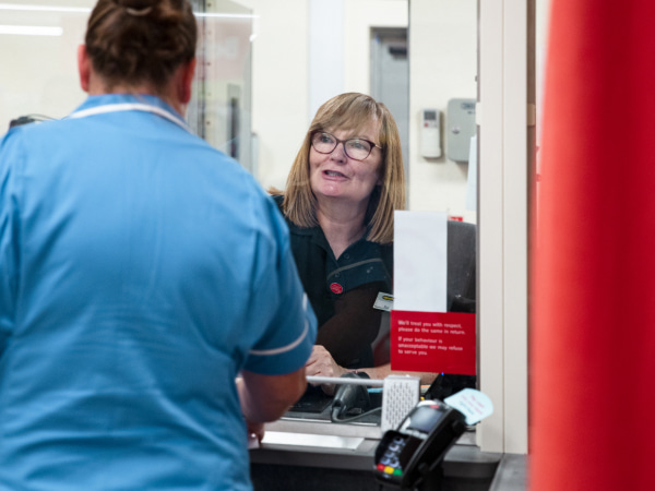 Woman in a nurse outfit being served by a female member of Post Office staff at a branch counter