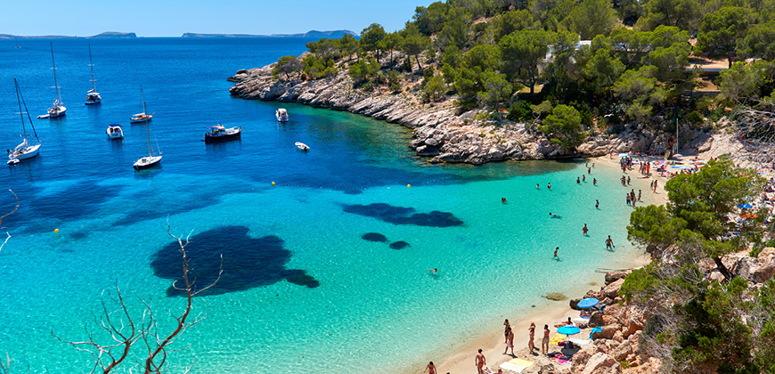 Rocky and green coastline with people on sandy beach surrounded with blue waters and boats